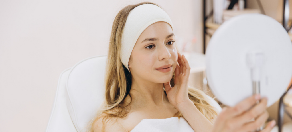 woman in a towel and headband examining her skin in a handheld mirror