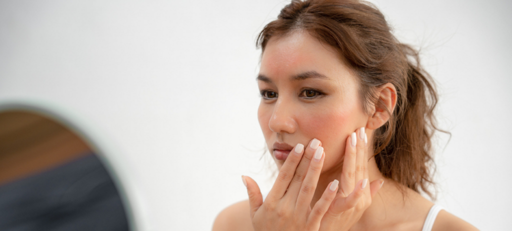 woman in a white tank top pressing skincare product into her cheeks at the mirror
