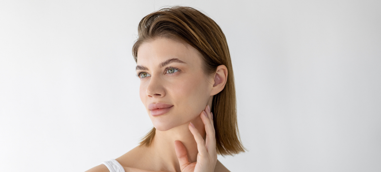 woman in a white tank top touching her jawline against a plain background